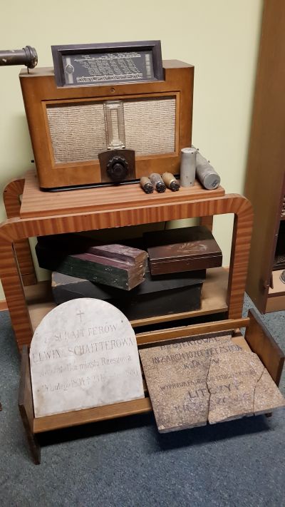 Prywatne radio Michała Franciszka Gottmana firmy Philips, przechowywane w czasie wojny  w sklepie, bardzo nowoczesne, z możliwością podłączenia do adapteru. Poniżej widoczna tablica nagrobna grobu rodziny Schaitter&oacute;w ze Starego Cmentarza oraz tablica ze sztucznego kamienia z informacją o autorze kamienicy, stanowiąca element wystroju elewacji lub klatki schodowej. Zdjęcie udostępnione przez Pana Grzegorza Dąbrowskiego